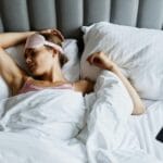 Kobieta śpi w łóżku z opaską na oczach, obok leży smartfon